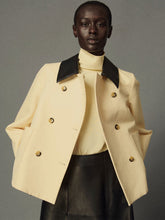 A model wearing a cream-colored double-breasted jacket with contrasting black lapels, paired with a cream turtleneck and a black leather skirt, set against a solid gray studio background. | HP - Fashion Clinic